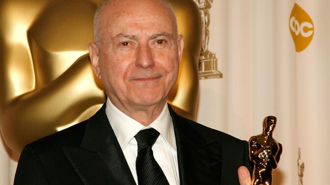 A man with light-tone skin holds an Oscar statuette. He is bald and wearing a tux with a black tie.