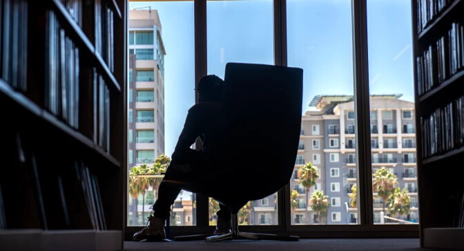 A man sits in one of the oversized chairs next to shelves of books inside a library.