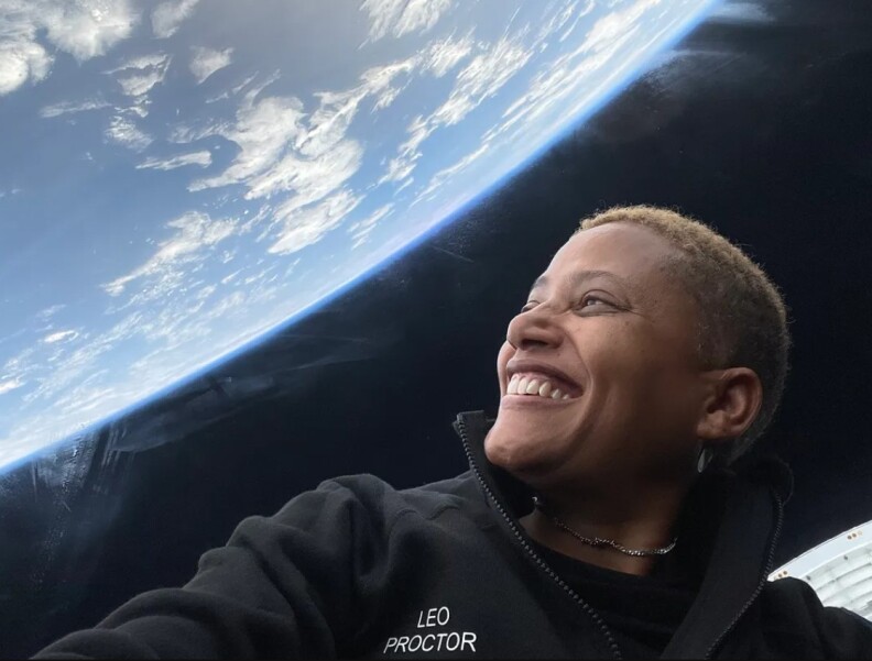 Black woman with short blonde hair smiles as she looks at the surface of earth from inside a space shuttle.