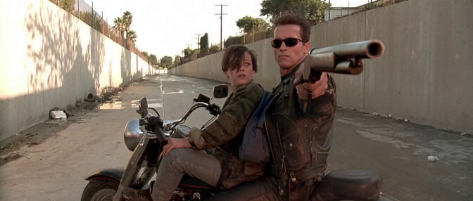 A young boy and a man wearing sunglasses and a leather jacket sit on a motorcycle, turned sideways in a flood-control channel. The man is pointing a rifle at something behind them while the boy looks at the man's face.