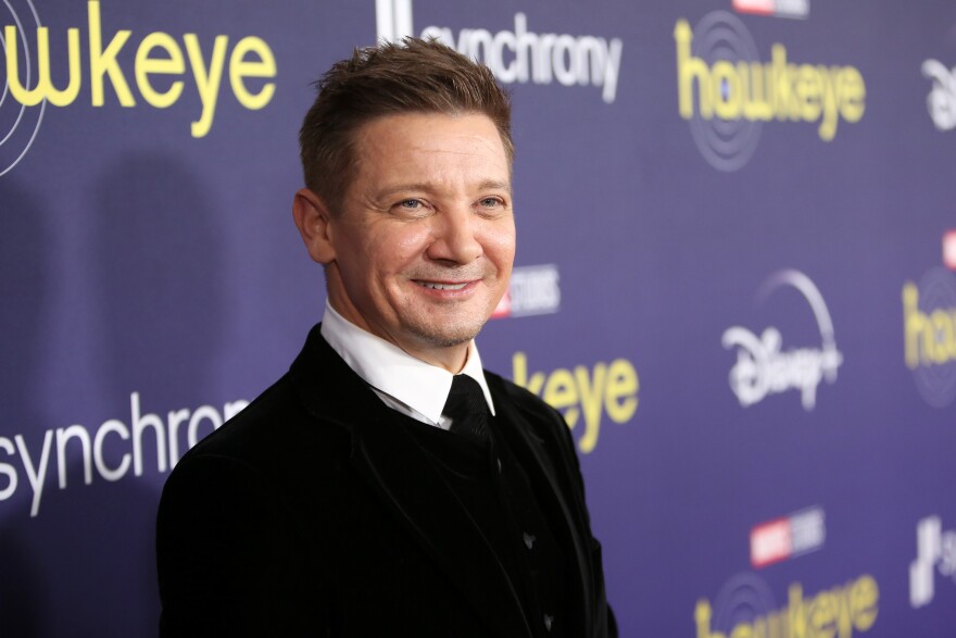 A white male with light skin tone and dressed in a black suit smiles for the camera at a premiere event. Behind him is a purple promotional background with the words "hawkeye" and "synchrony" on it.