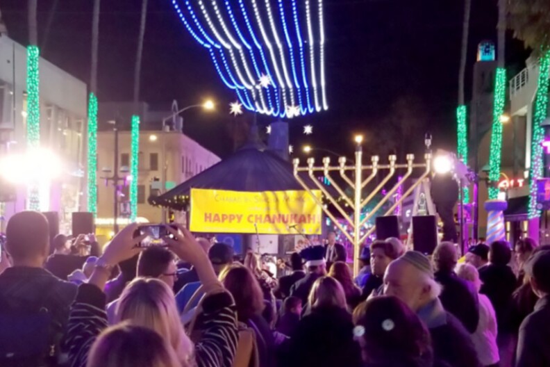 A crowd of people look on at a stage with a giant menorah. A yellow sign reads "Happy Chanukah!"