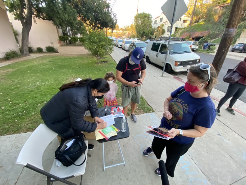 Three adults gather around a small folding table on a sidewalk. One adult is bent over the table writing on a sheet of paper as the other two adults look on.
