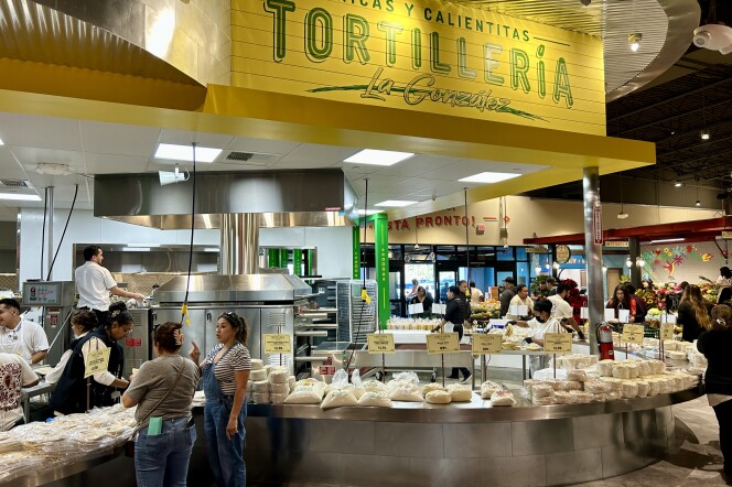 A wide shot of a long curving counter stacked with tortillas and masa for making tortillas. There are customers standing around the outside, and workers manning large metal machines on the inside. Above the area a bright yellow sign reads "Tortillería La González."