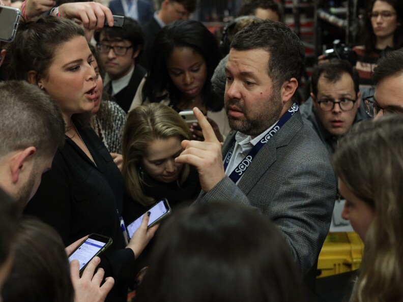 People, many looking at their cell phones, crowd close together.