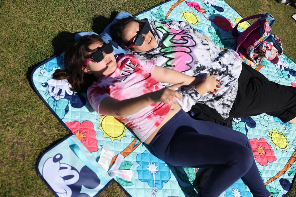 Two little girls wearing colorful shirts and pants lay on a Mickey Mouse blanket while looking up at the sky and wearing eclipse glasses. 