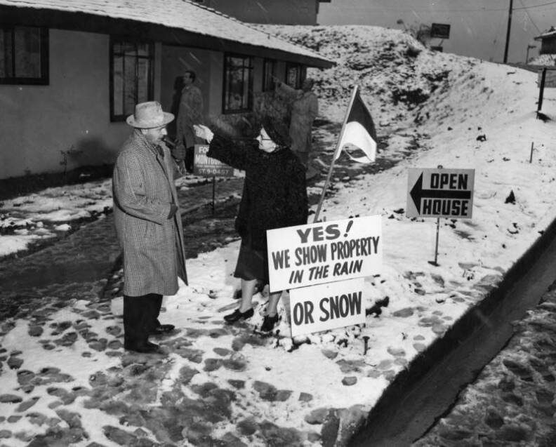 Two people with light-tone skin wear coats and hats standing in a lawn covered in snow. An open house sign points to the entry next to another sign reading: "Yes! We show property in the rain" with a sign added below: "or snow."
