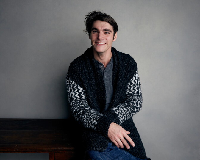 RJ Mitte, a smiling person with dark, tousled hair, sits against a gray studio wall, looking off to the side. They wear a black-and-white patterned knit cardigan over a gray henley.