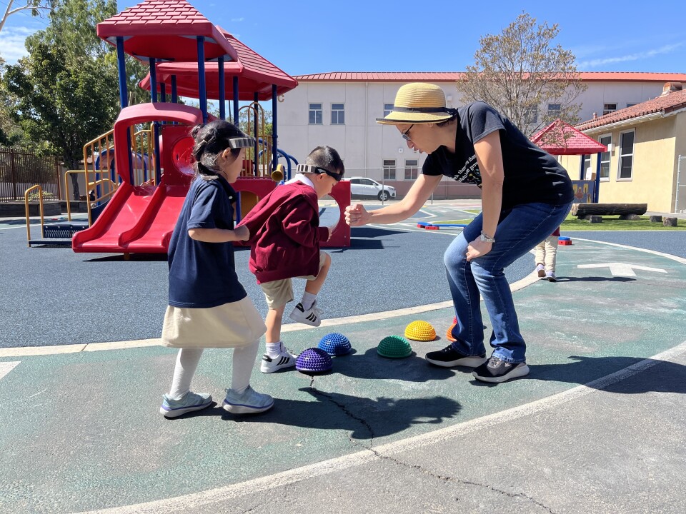 Two young children step on colorful balancing toys as a woman with a hat and black T-shirt holds out a fist for them to fist pump. 
