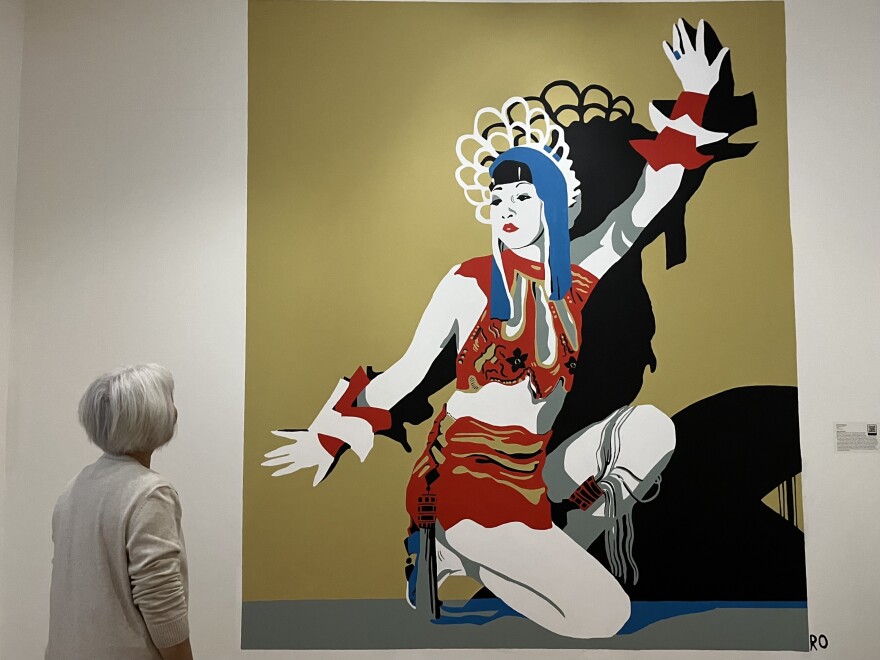 A white hair-bobbed woman in a white sweater looks at a 12-foot-tall mural of Anna May Wong inside a museum.