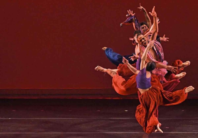 Dancers in orange parachute pants form a vertical line on a stage with their bodies, feet and arms are extended in a dance move.