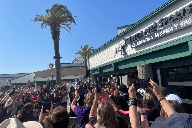 A crowd holds their phones up outside a bar. The words "Watch Me! Sports" and "Celebrating women's sports" are visible on the sign.