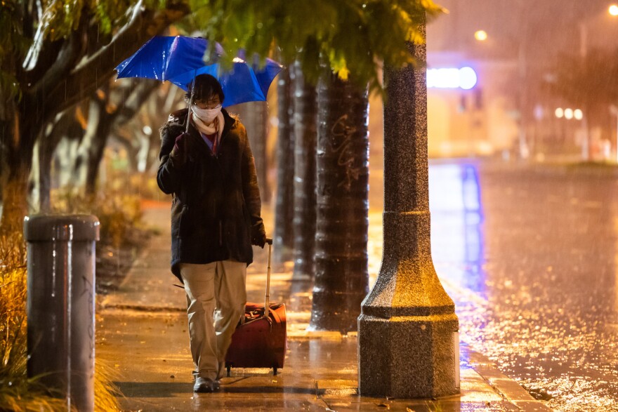 A woman wearing a black rain coat, tan pants, and a dragging a red rolling suitcase, holds a blue umbrella as she walks on a rainy sidewalk. Next to her, the road is empty and full of rain.