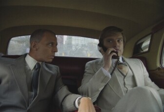 Two men in suits sit in the back of a car. the man on the right holds an old cellphone to his ear while the man on the left leans over to listen. 