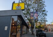 The photo shows the exterior of a Philz Coffee shop, easily identifiable by a large yellow sign with the Philz Coffee logo. The shop features large windows, allowing a view inside where you can see hanging lights and tables.