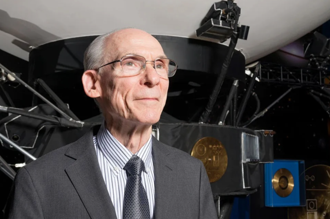 An older adult man stands in front a space craft wearing a great suit change, striped shirt, and grey tie. The man has glasses and is looking past the camera. 
