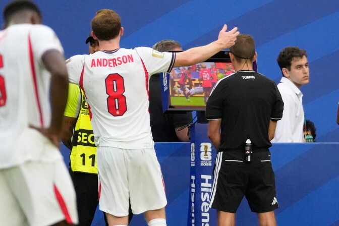 A referee watches a monitor as players in a white and red jersey wait and react.