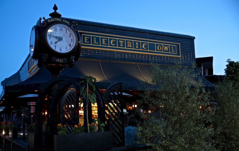 The exterior of Electric Owl, a restaurant and bar with a dark tiled facade and a large vintage-style street clock out front. The sign above the entrance glows with the name “Electric Owl” in gold letters, while the warm lights of the bar can be seen inside at dusk.