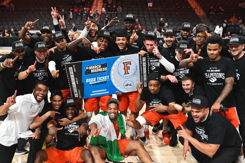 Cal State Fullerton basketball players gather for a photo, dressed in black t-shirts and orange shorts. Two of them hold a blown-up ticket to March Madness.