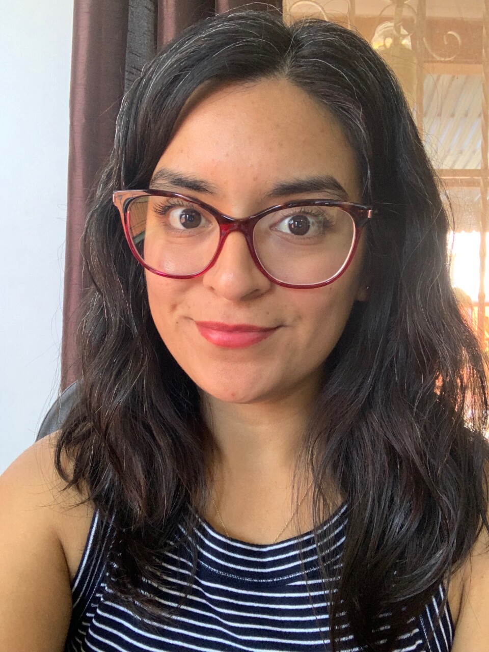 An image of a woman's face. She wears red-rimmed glasses, a striped black-and-white sleeveless shirt, and has dark hair. 