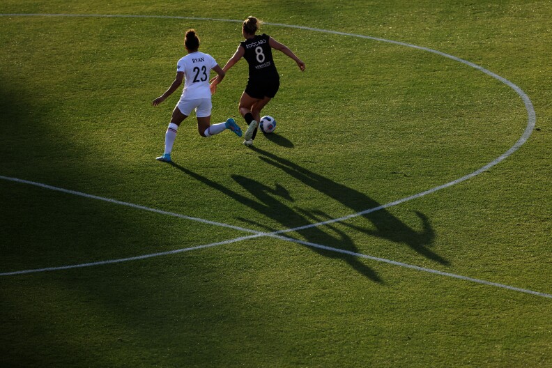Two soccer players chase after a ball in the middle of the field, their long shadows stretched across the grass in the slanting sunlight.