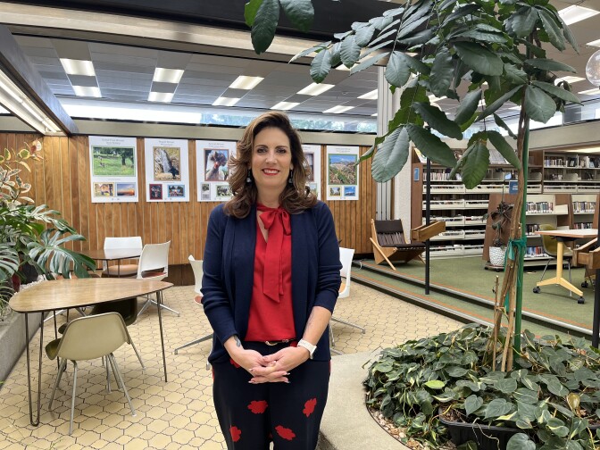 A white woman in a red blouse and blue jacket poses by chairs and tables in a library.