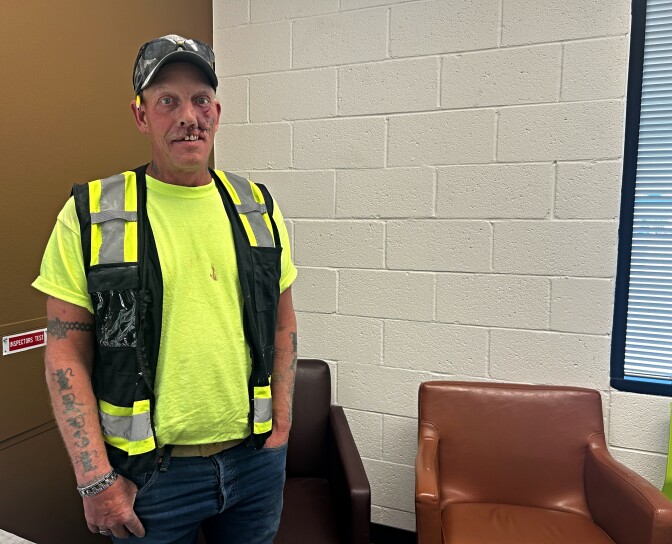 A man wearing a neon yellow T-shirt and reflective vest stands with his hands in his pockets. 