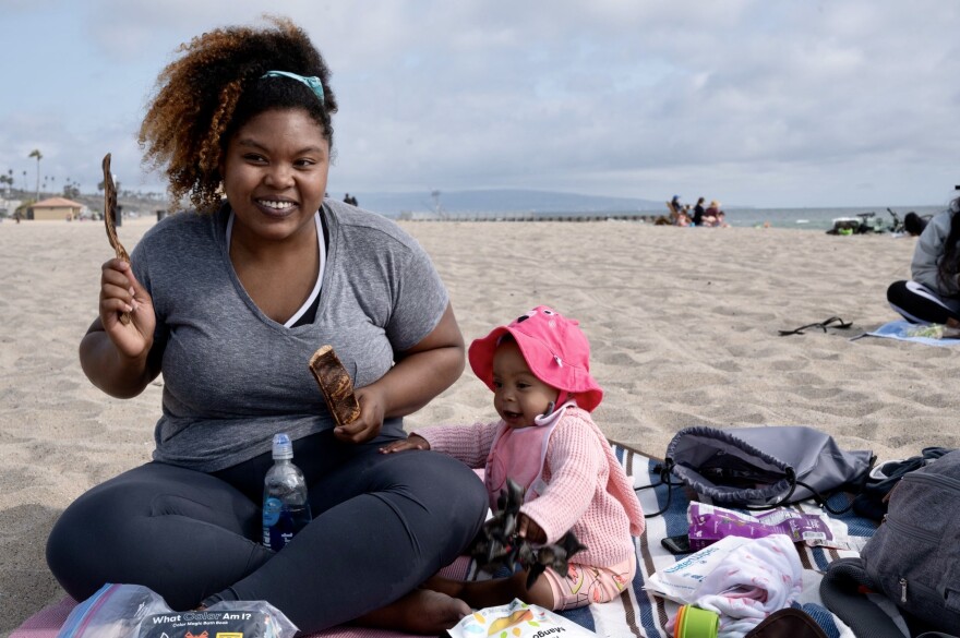 A Black mother with a huge smile sits cross legged on a blanket on the beach. Her baby, dressed in a pink outfit, complete with a magenta sunhat to match, sits next to her with her hand on her mother's lap. The mother holds pieces of palm fronts like drumsticks in the air, as if playing a game.