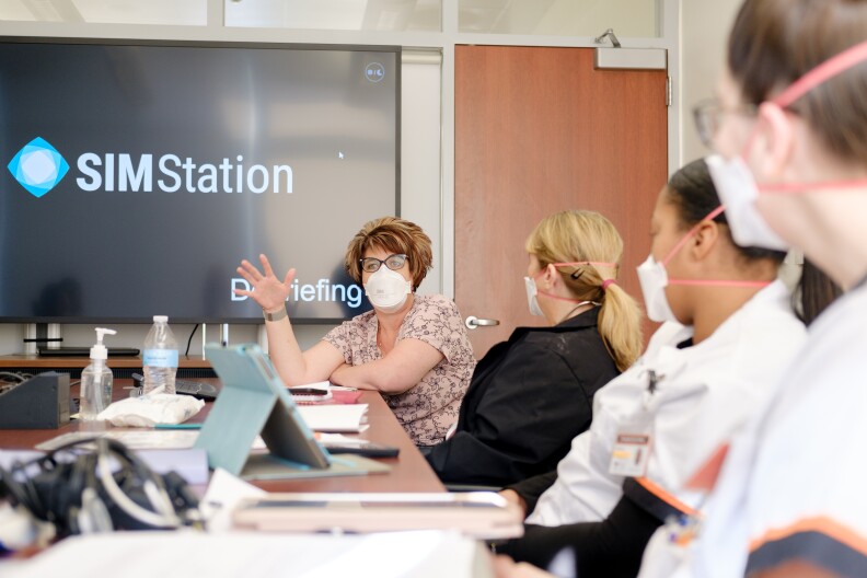 Four women sit at a conference table, all wearing masks. Three of the women turn to look at the one at the end of the table as she speaks. A large TV in the background displays the word "SIMstation."