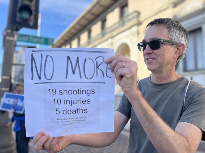 A person holding a white sheet of paper with bold, handwritten and printed text. The paper reads:
At the top, in large handwritten letters: “NO MORE” Below that, in printed text:
“19 shootings 10 injuries 5 deaths”