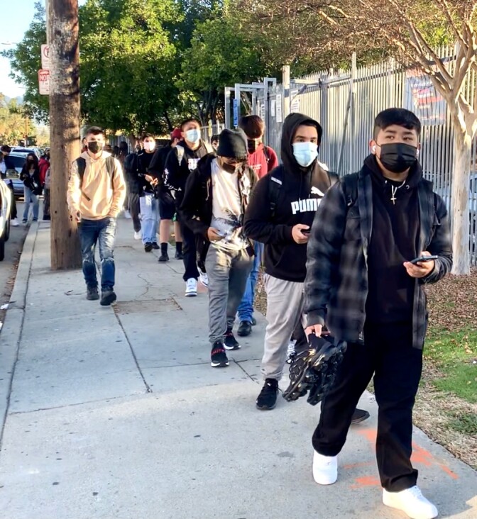 Male high school students in hoodies and joggers stand in line on a sidewalk outside their high school.