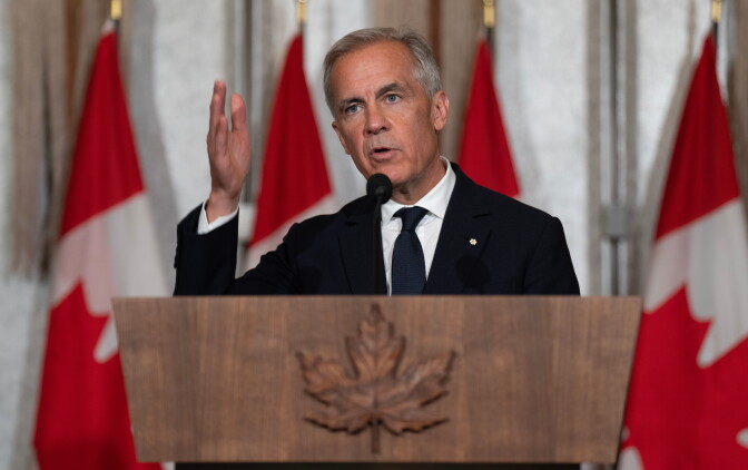 Canada Prime Minister Mark Carney, a man with light skin tone, wearing a black suit and tie, speaks behind a podium with a wooden leaf on it as he stands in front of Canadian flags.