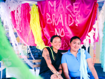 Two women sit infront of a hot pink satin fabric that says "Make braids, no raids." The fabric is is surrounded by colorful strings of lace and ribbon.