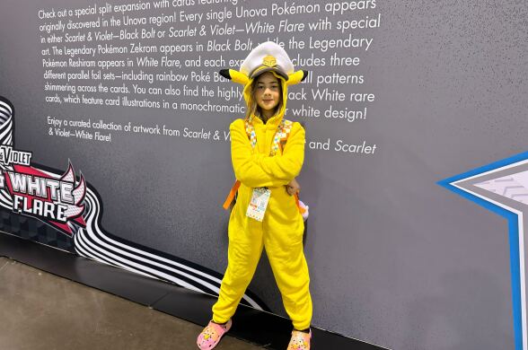 A young girl with light skin tone, in a bright yellow full-body onesie, stands for a portrait. Her arms are crossed, with confidence.