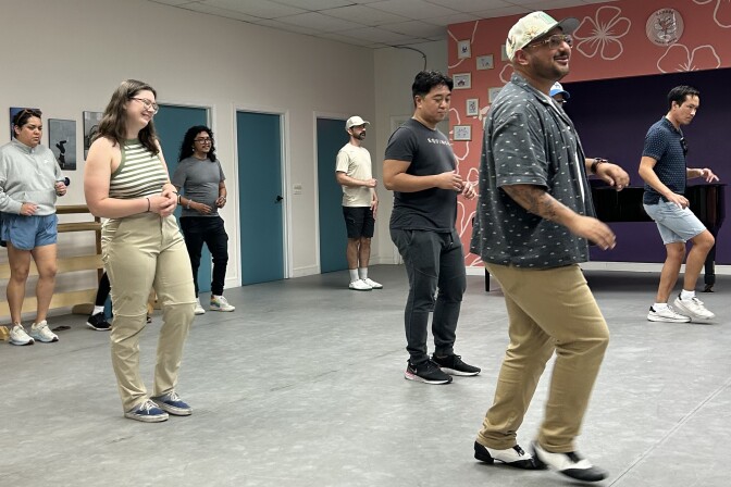 A dance studio with a handful of people spread out. At the front of the room is an instructor wearing glasses, a tan cap and a navy blue button-up shirt. 