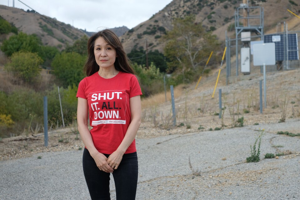 A middle-aged Asian woman with light skin tone and long straight brown hair poses for a photo. She wears a red shirt that says "SHUT IT ALL DOWN" and black pants. Rolling hills are behind her. 