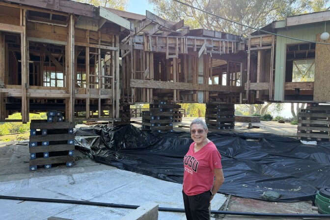 A woman, with cropped grey hair and wearing a red tshirt and black pants, stands in front of a hollowed out home raised on wooden planks. 