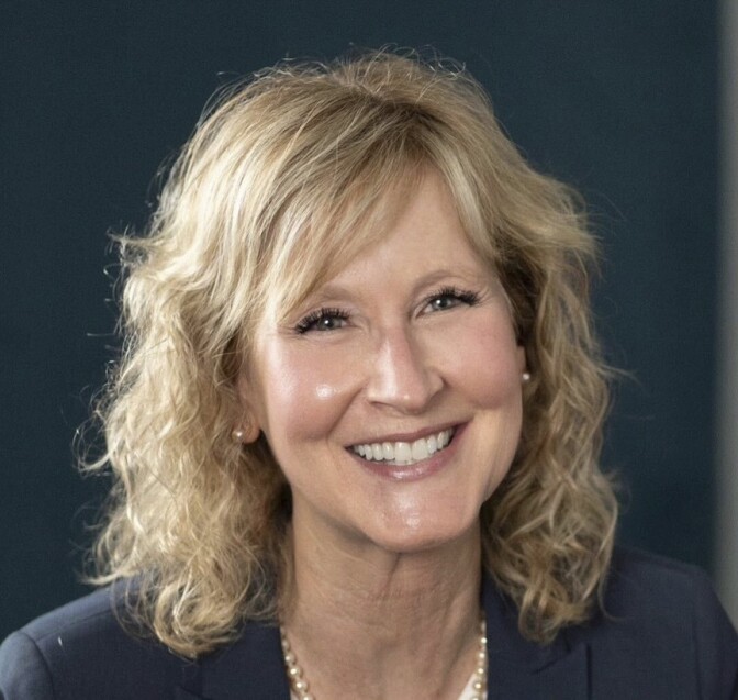A woman with blonde, shoulder length hair, smiles while seated in front of a black background wearing a black blazer 