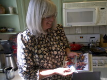 A light skinned woman standing in her kitchen smiles as she holds a photo album.