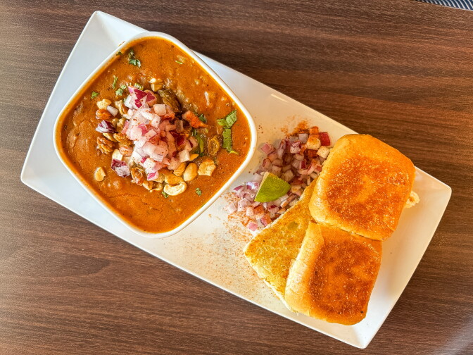 An overhead shot of the Honest Special Bhaji Pav — a rich orange-red spiced curry topped with cashews, raisins, and diced red onion in a white bowl, served alongside two golden toasted pav rolls on a white rectangular plate.