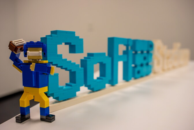 A Lego version of one of the Rams' players, in yellow and blue, holds a Lego football in front of a SoFi Stadium sign made of Lego.
