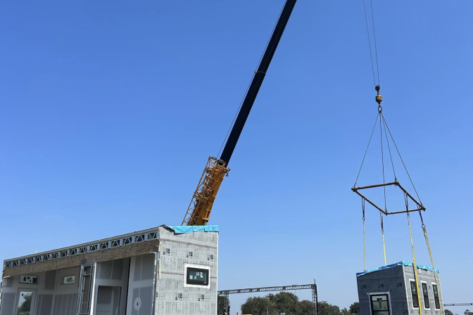 Students stand below a long crane machine, cradling the small portion of a house in pieces. The main portion of the house is at the front and tho the left of them. They are all wearing hard hats and safety vests.
