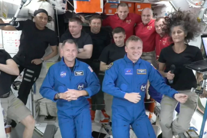 Two man are seen in blue space suits in front of eight other people behind them. They appear to be floating without gravity with gear and wires seen around them. 