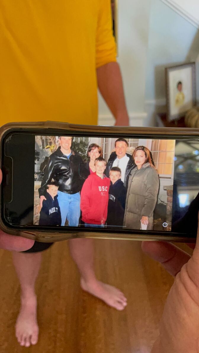 A photo of a photo on an Iphone showing the Leslie family with Brad Pitt and Angelina Jolie. You can see part of a hand holding the phone and in the background, behind the phone, is a man standing barefoot on a wooden floor, wearing black shorts and a long-sleeve yellow t-shirt.