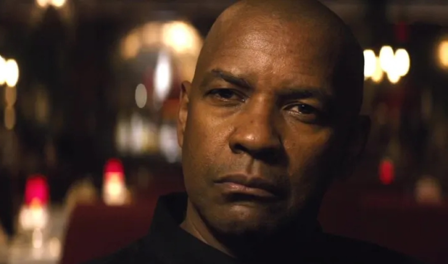 A close up on Denzel Washington's face, only the neck up visible. The black collar of his button up can be seen. Behind him, out of focus, is a dimly lit restaurant with red booths and red candle holders on the tables. Washington looks intensely at someone sitting in front of him out of frame. His eyes are slightly narrowed. 