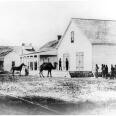 A black and white view of the U.S. Army Headquarters at Wilmington. The building is of wood and has large doors. In front of the building is a camel with two humps, a person holding the reins of a horse, a group of people standing in the front yard.