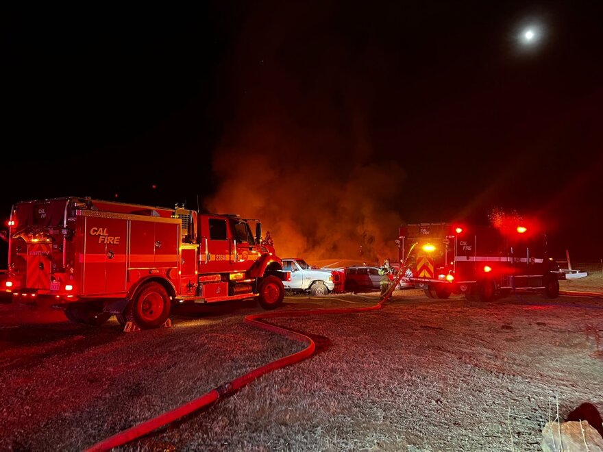 Two fire trucks are shown near a car at nighttime. Their red lights are on. Smoke can be seen in the background.