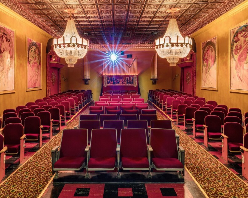 A view from the stage of a small old movie theater with red velvet chairs, two chandeliers, organ pipes in the background and art deco paintings on the walls.