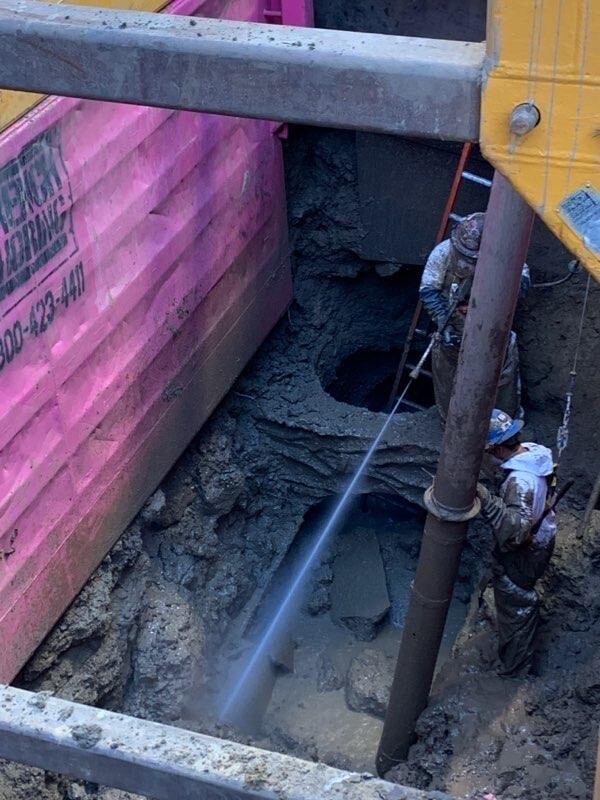 Two sanitation workers stand in a dirt pit next to a pipe and pink retaining wall. One worker sprays a stream of water deeper into the pit.