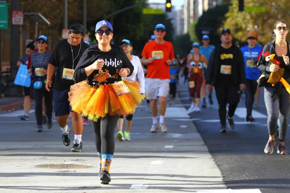 A light-skinned woman wearing a blue Dodgers hat and an orange and yellow tutu runs on a street. Behind her, several other people run and walk in a race. 